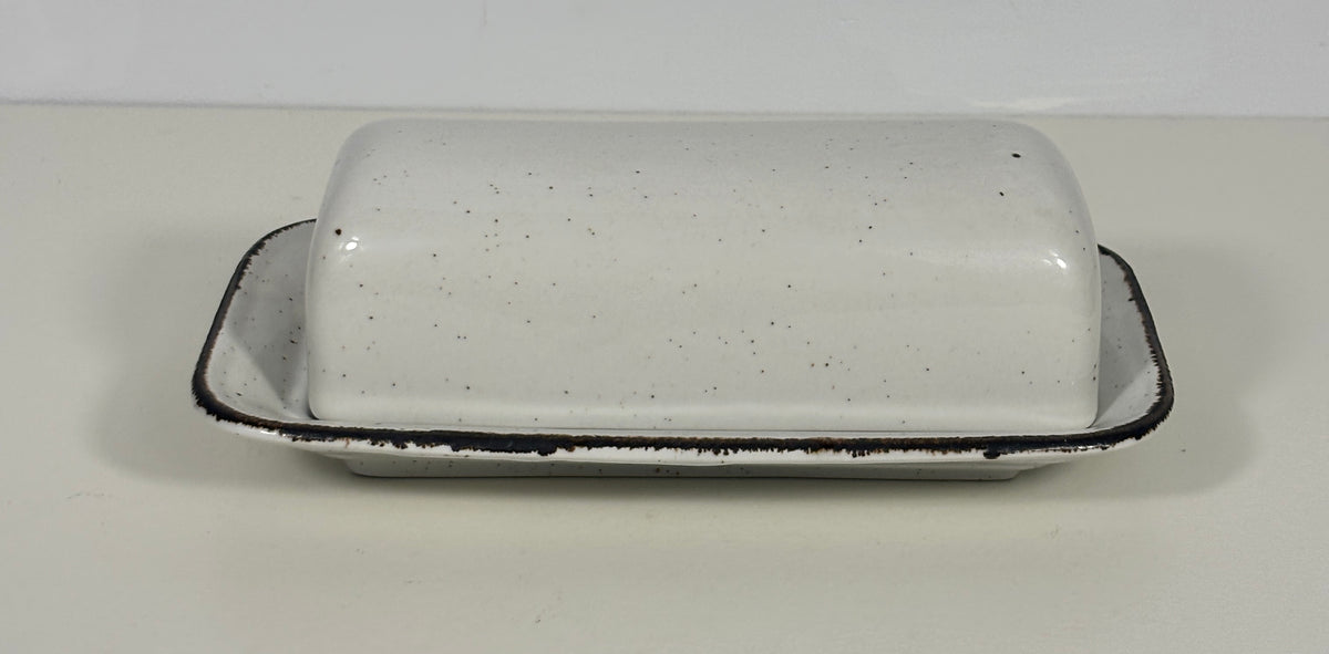 A white speckled ceramic butter dish with a matching lid, featuring a rustic black rim, set against a plain off-white background.