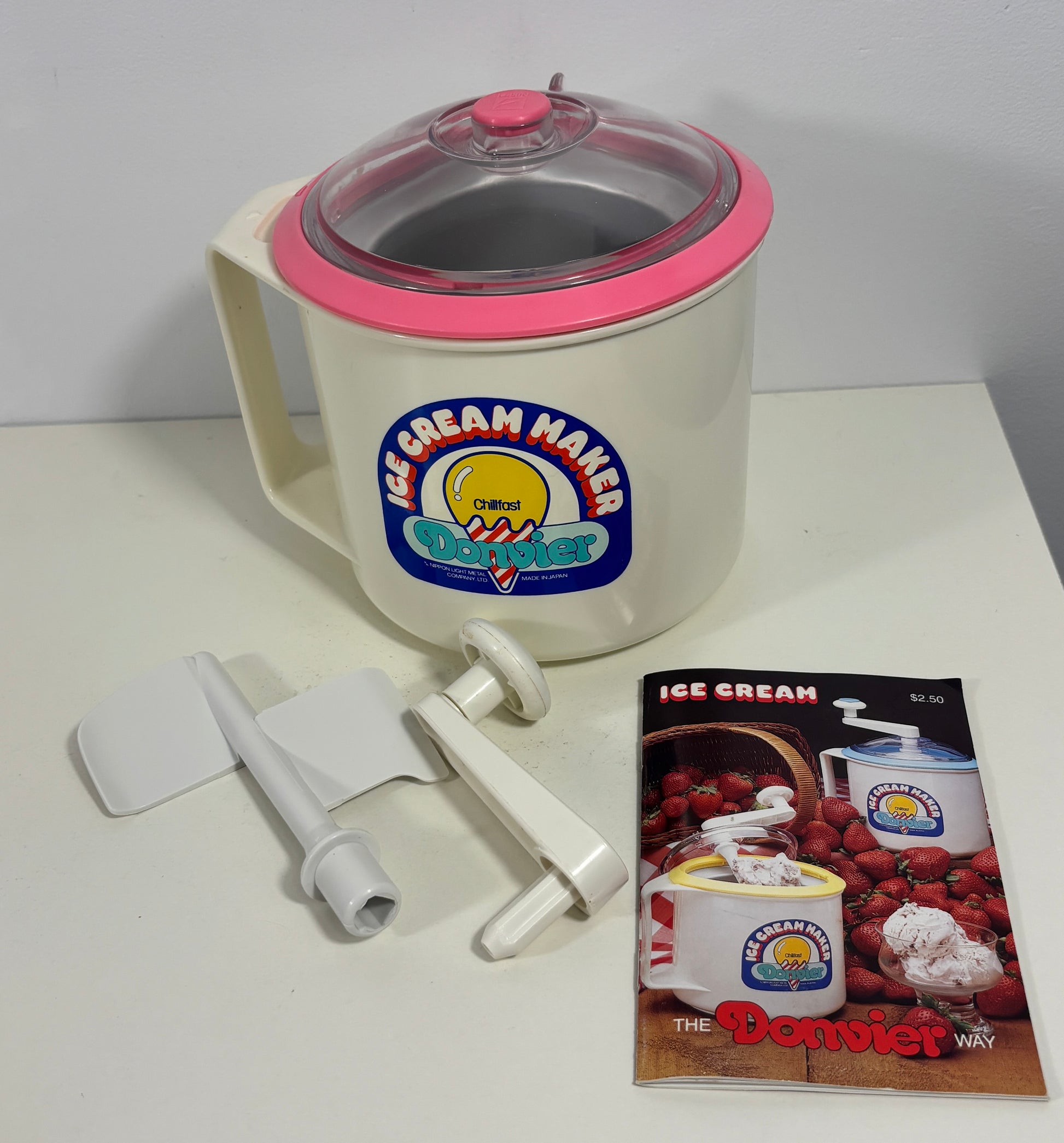 A vintage Danviet ice cream maker with a cream-colored body, pink trim, and clear lid, shown with three white plastic attachments and a small printed booklet on a white tabletop against a light gray wall.