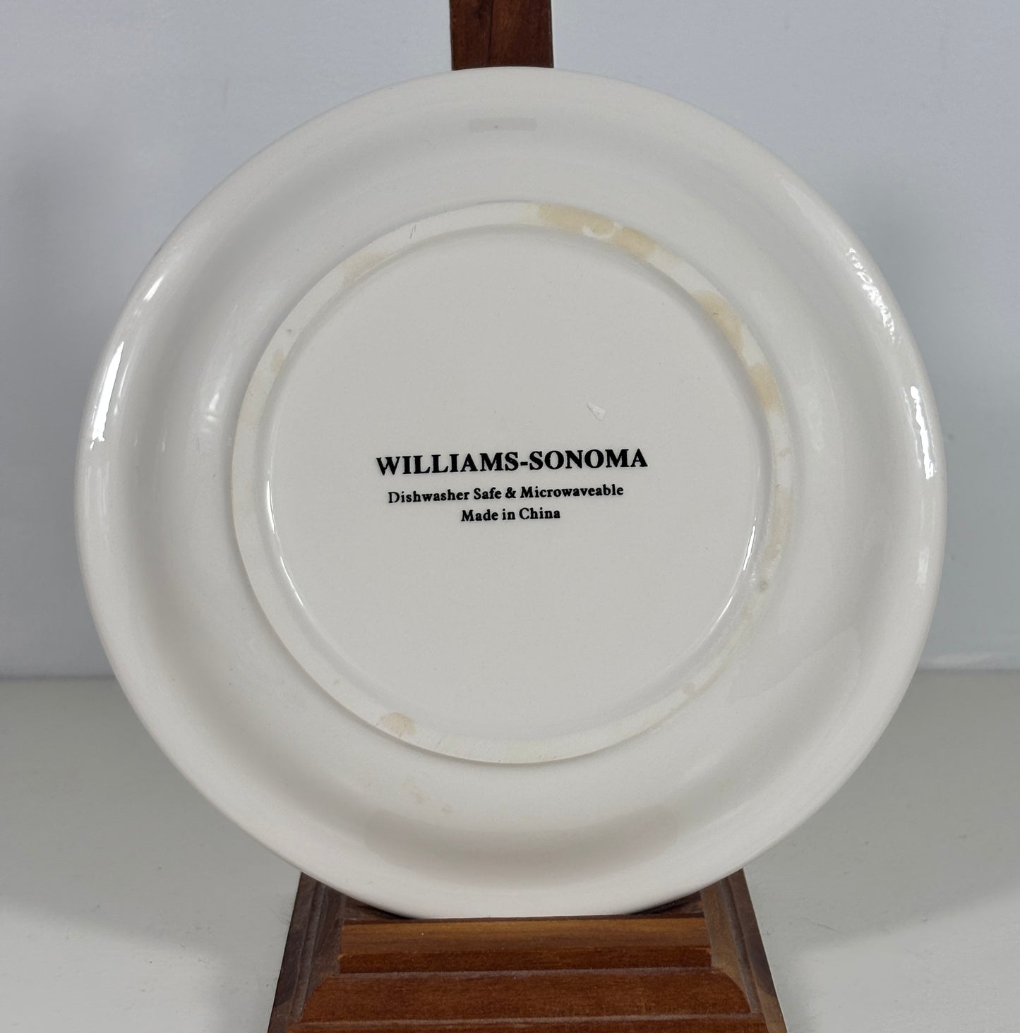 A white ceramic plate shown from the back, resting on a wooden display stand against a plain light background. The plate features black printed text in the center reading 'WILLIAMS-SONOMA Dishwasher Safe & Microwaveable Made in China'.