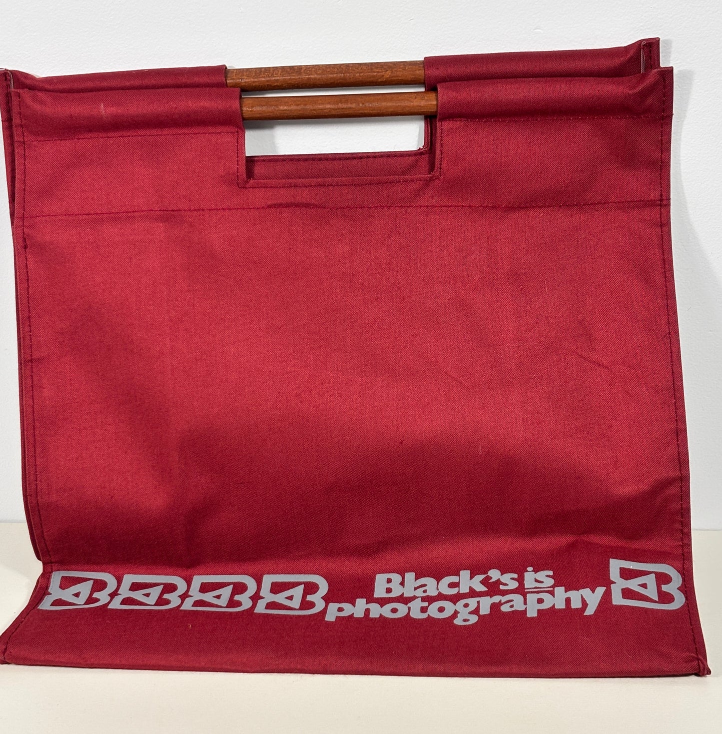 A dark red fabric tote bag with a wooden handle, featuring the white printed text 'Black's is photography' and a stylized 'B' logo along the bottom edge.