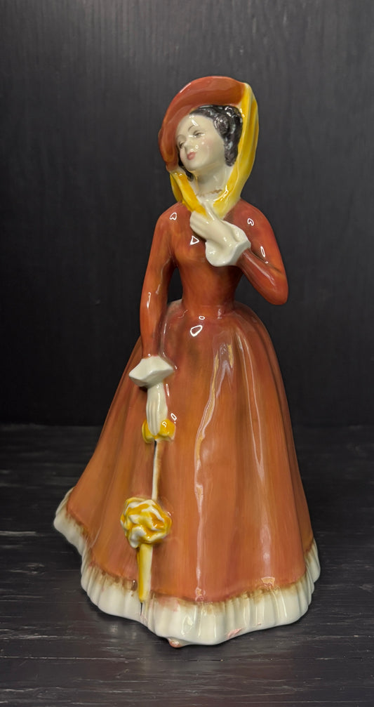Vintage ceramic figurine of a woman in a brown dress with a yellow headscarf and basket, on a dark background.