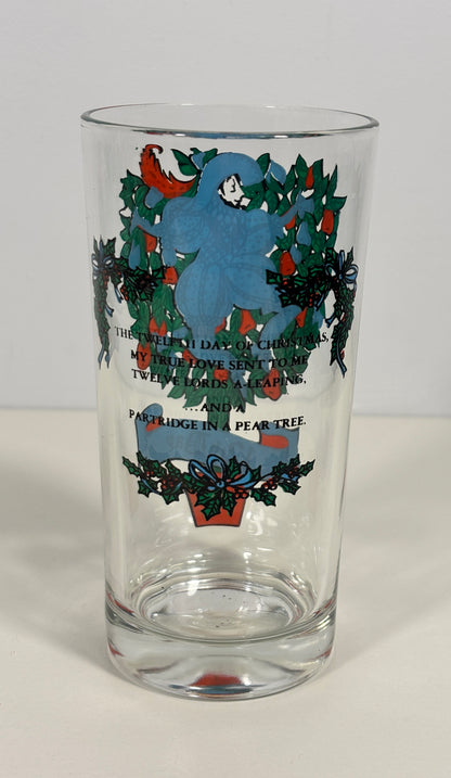 A clear glass pint glass featuring a Christmas-themed illustration of a blue bird, green holly leaves, red berries, and black text from 'The Christmas Carol' on a white background.