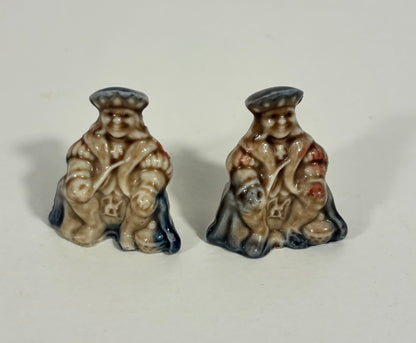 A pair of small ceramic figurines depicting seated figures in vintage clothing, shown on a plain off-white background.