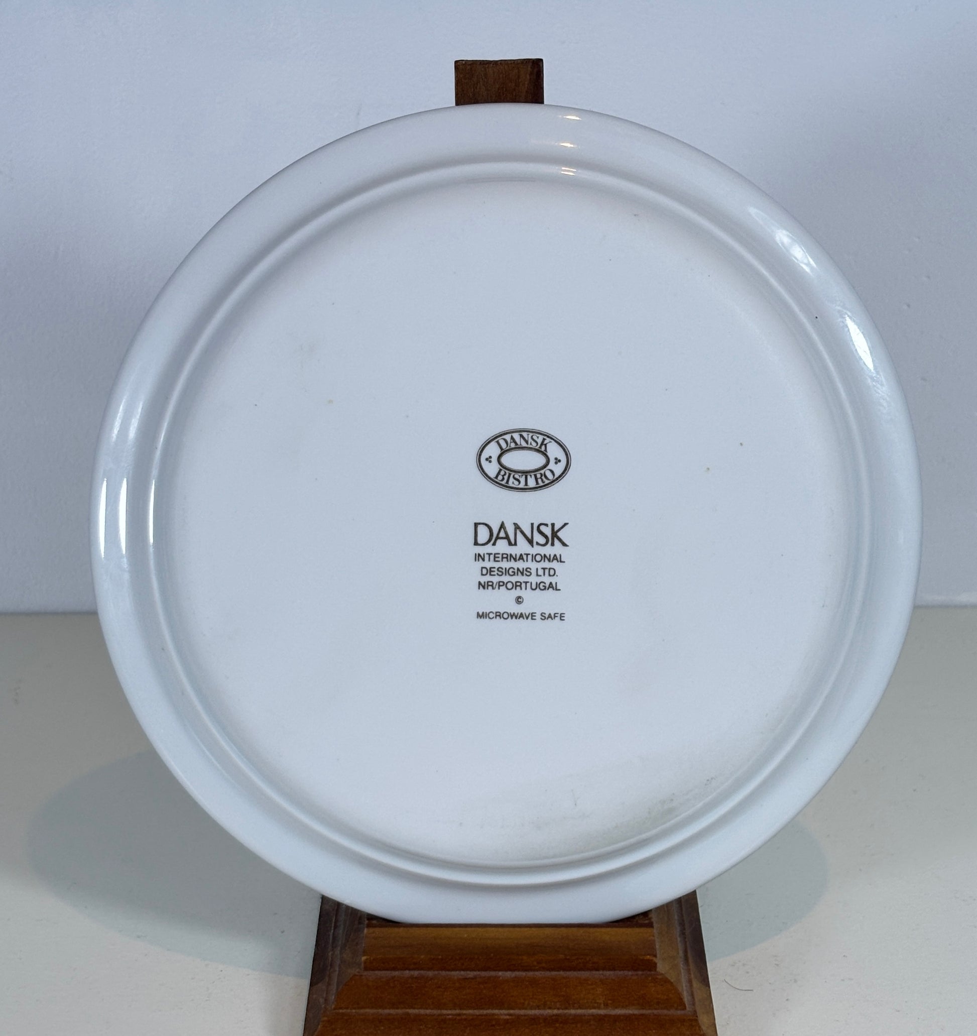A white ceramic Dansk International plate shown from the back, resting on a small wooden display stand against a plain light background.