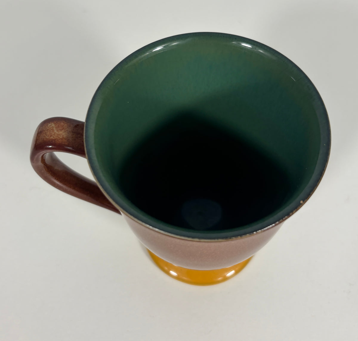 A ceramic mug with a dark green glazed interior, a brown exterior and handle, and a small orange base, shown from a top-down angle on a white background.