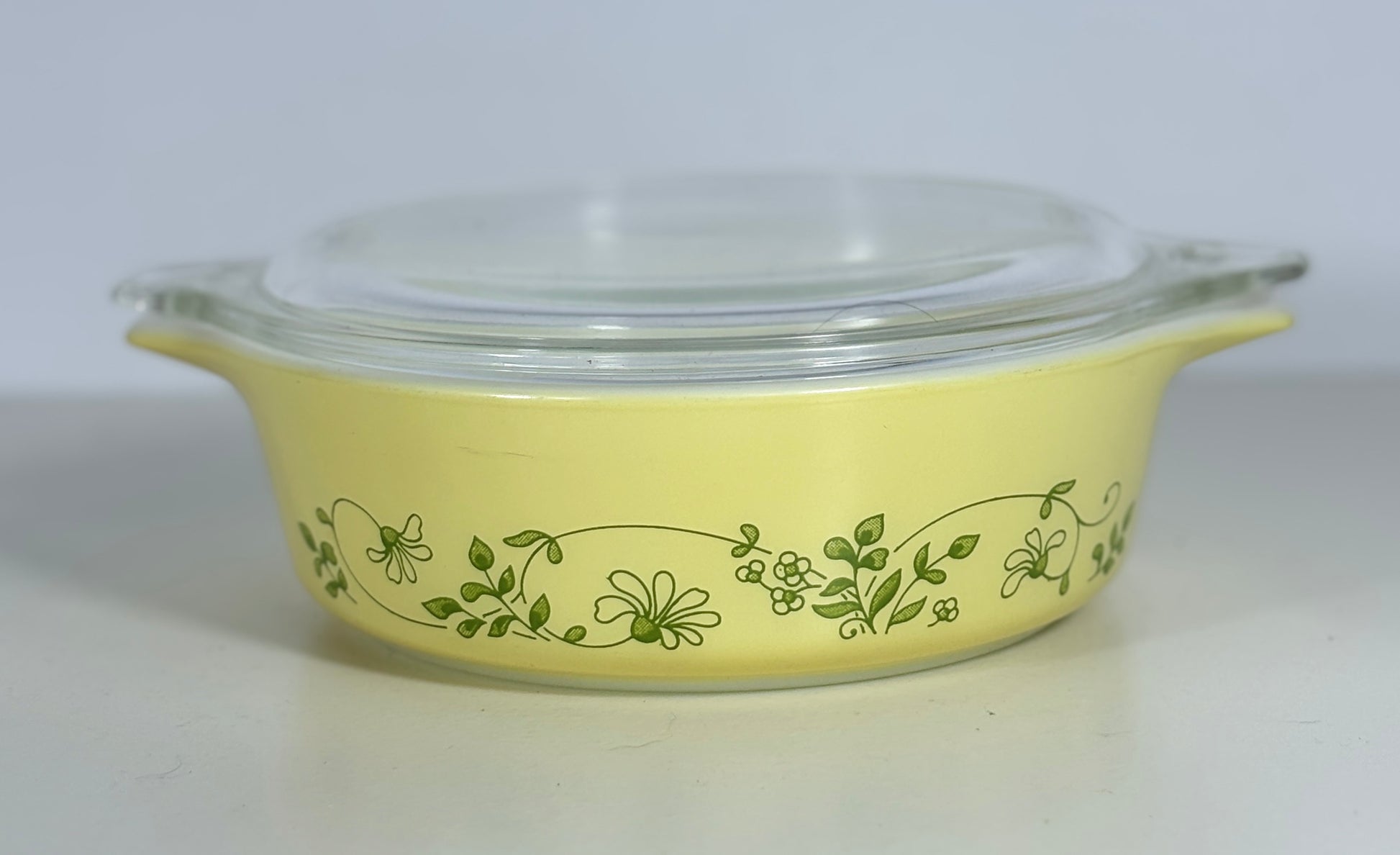 A yellow glass casserole dish with a clear glass lid and green floral vine pattern, photographed on a plain light background.