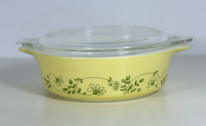 A yellow glass casserole dish with a clear glass lid and green floral vine pattern, photographed on a plain light background.