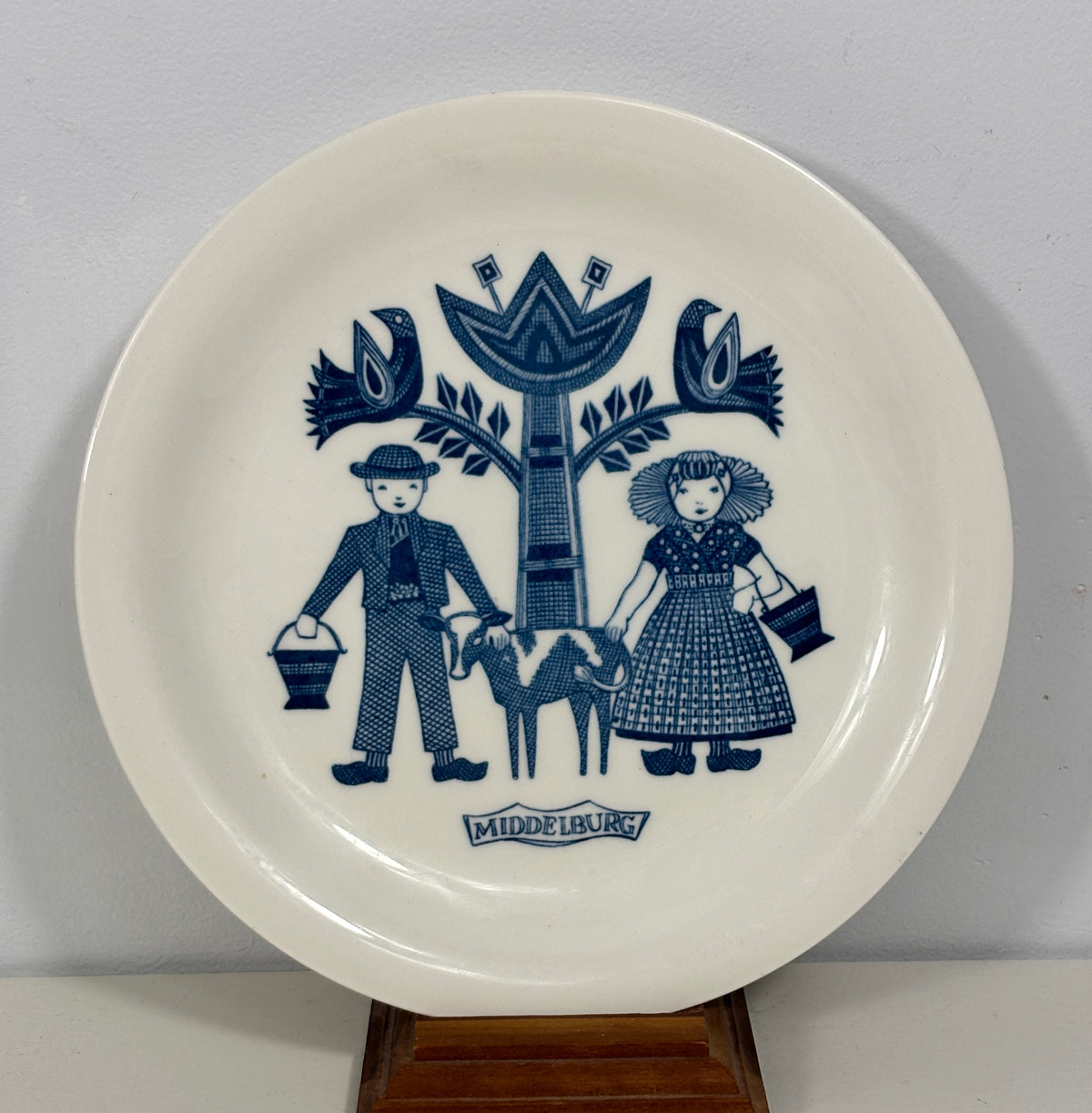 A white ceramic decorative plate with a blue Delft-style illustration of a man, woman, and goat under a tree, with the brand name 'MIDDELBURG' printed at the bottom, displayed on a wooden stand against a light gray wall.