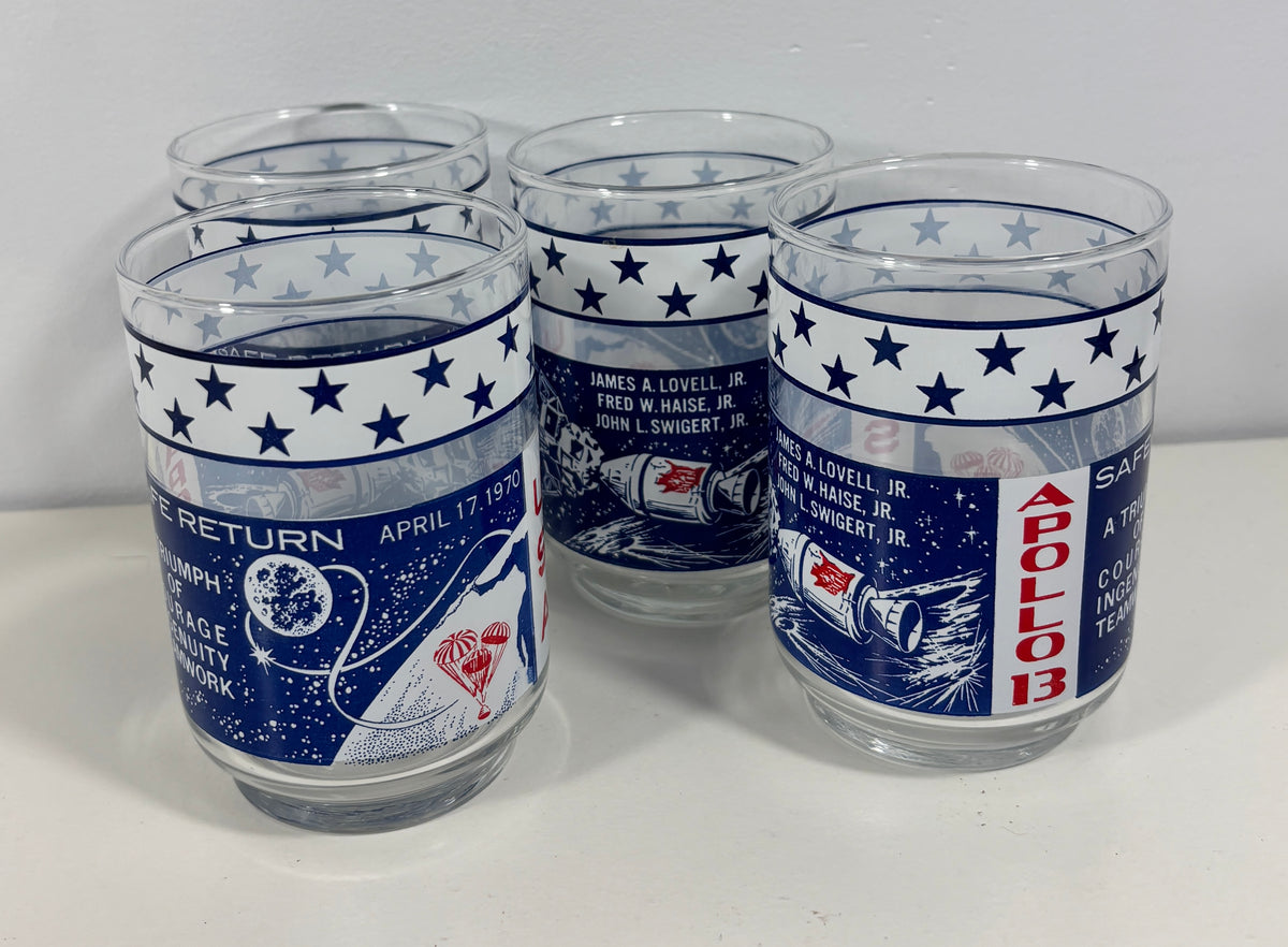Five clear glass tumblers with blue and white Apollo 11 space-themed printed bands, including stars, text, and a spacecraft illustration, photographed on a white surface against a white background.
