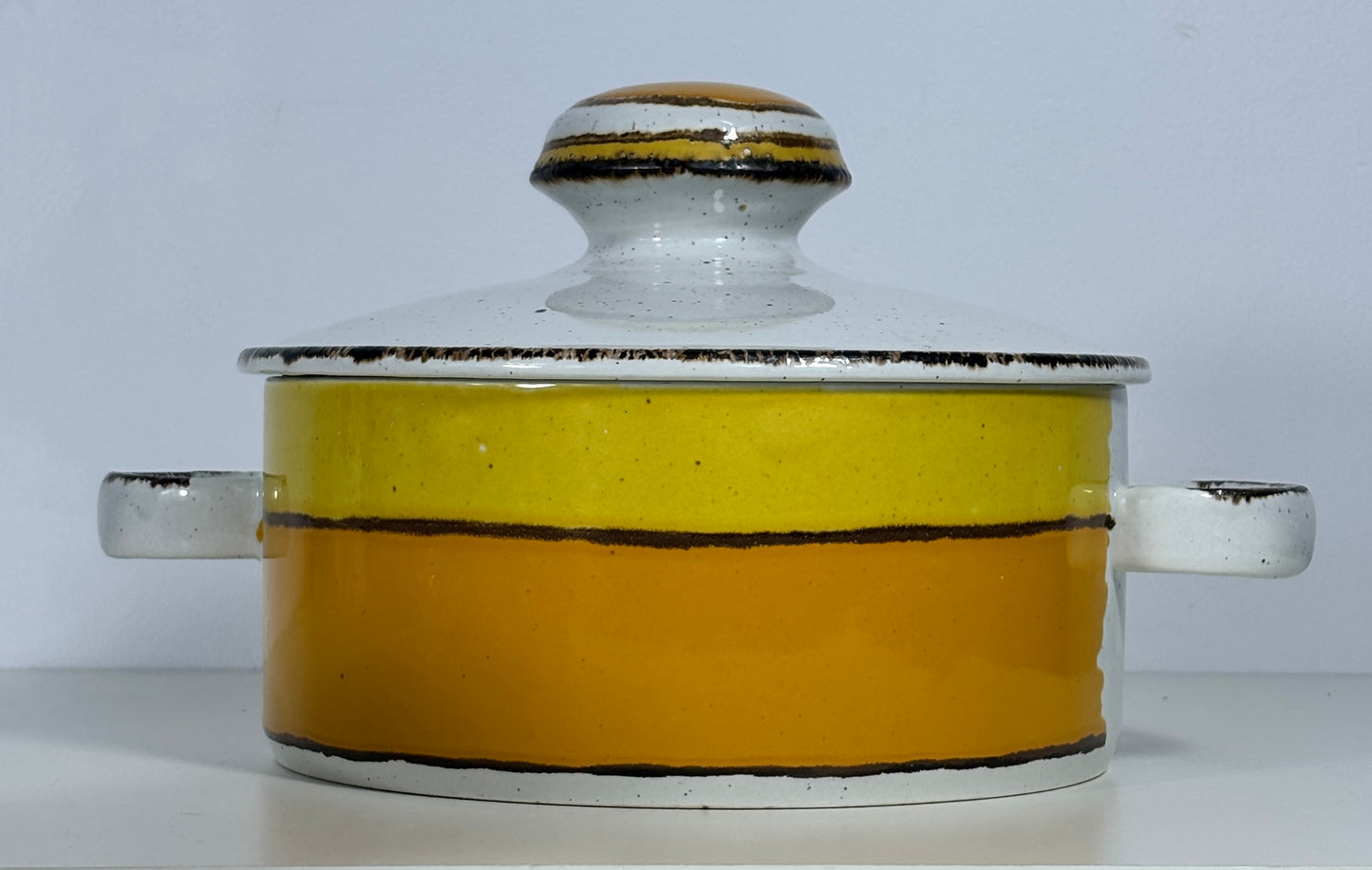 A ceramic lidded casserole dish with a two-tone yellow and orange body, white handles, and a white glazed lid with a knob, set against a plain light gray background.