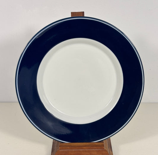 A round ceramic plate with a wide dark navy blue rim and a white center, displayed on a small wooden stand against a plain light background.