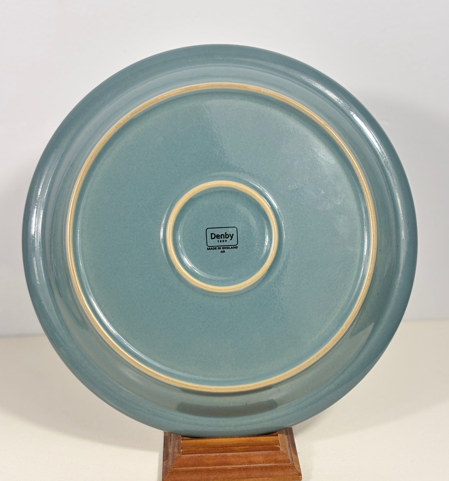 A round ceramic plate with a dusty teal-blue glaze and a cream-colored double rim, displayed on a small wooden stand. The inside of the plate features a central logo that reads 'Derby Creamware Co.