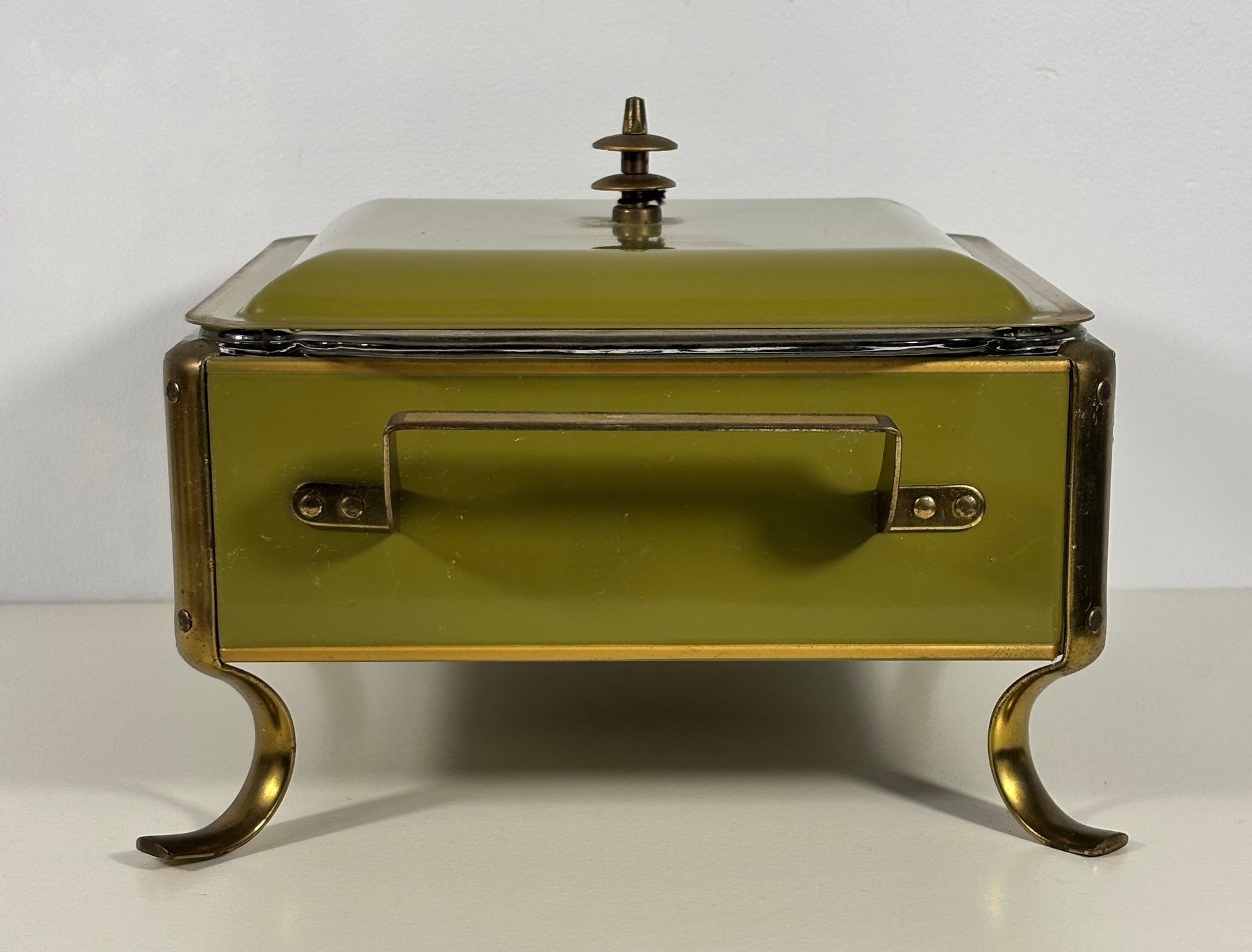 An antique-style lidded serving box or chafing dish with a muted olive green enamel finish and brass-toned metal legs, handle, and trim, set against a plain white background.