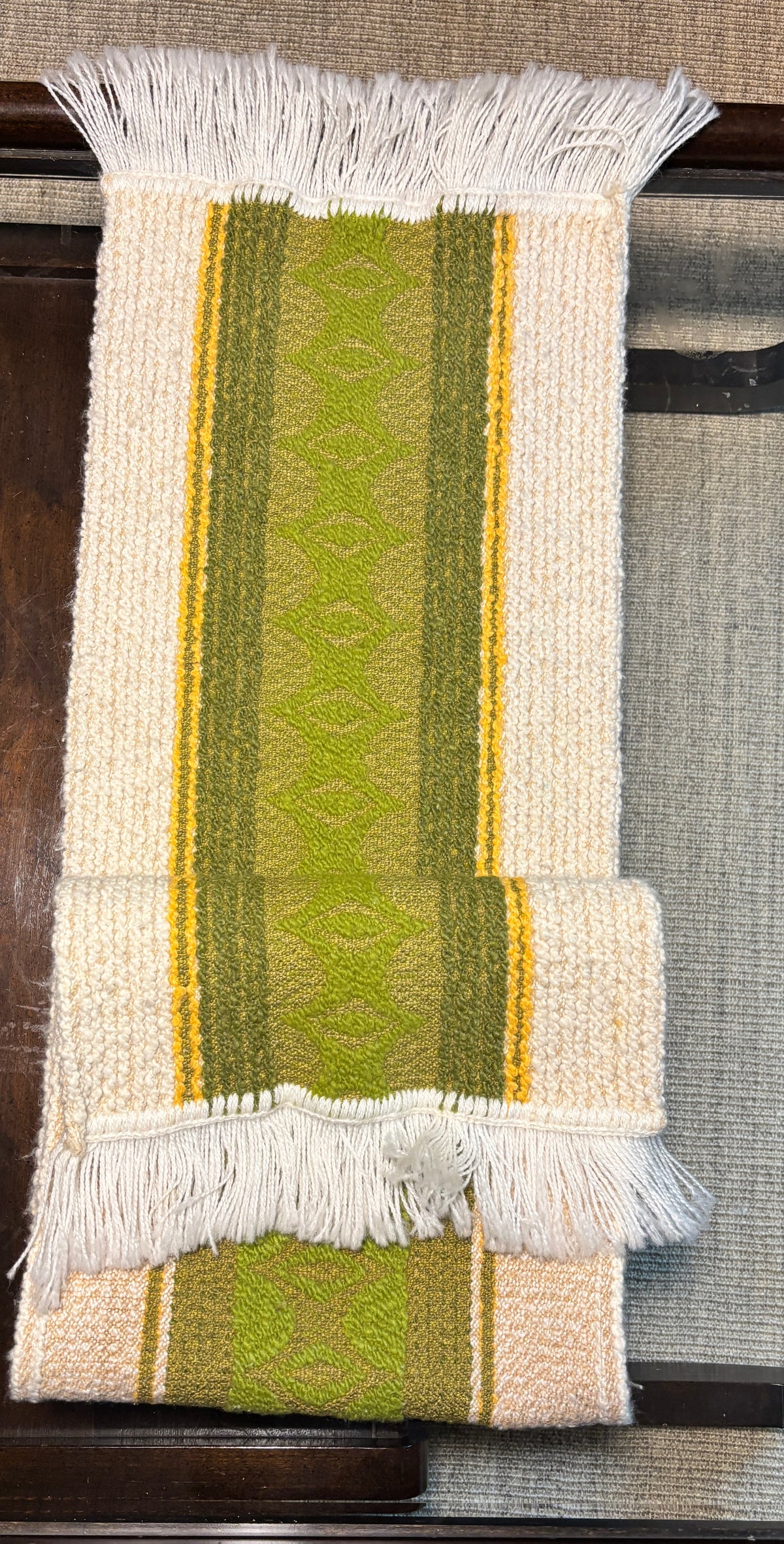 A folded woven textile runner or throw with white fringe, featuring vertical stripes in cream, olive green, and mustard yellow on a dark wooden surface.
