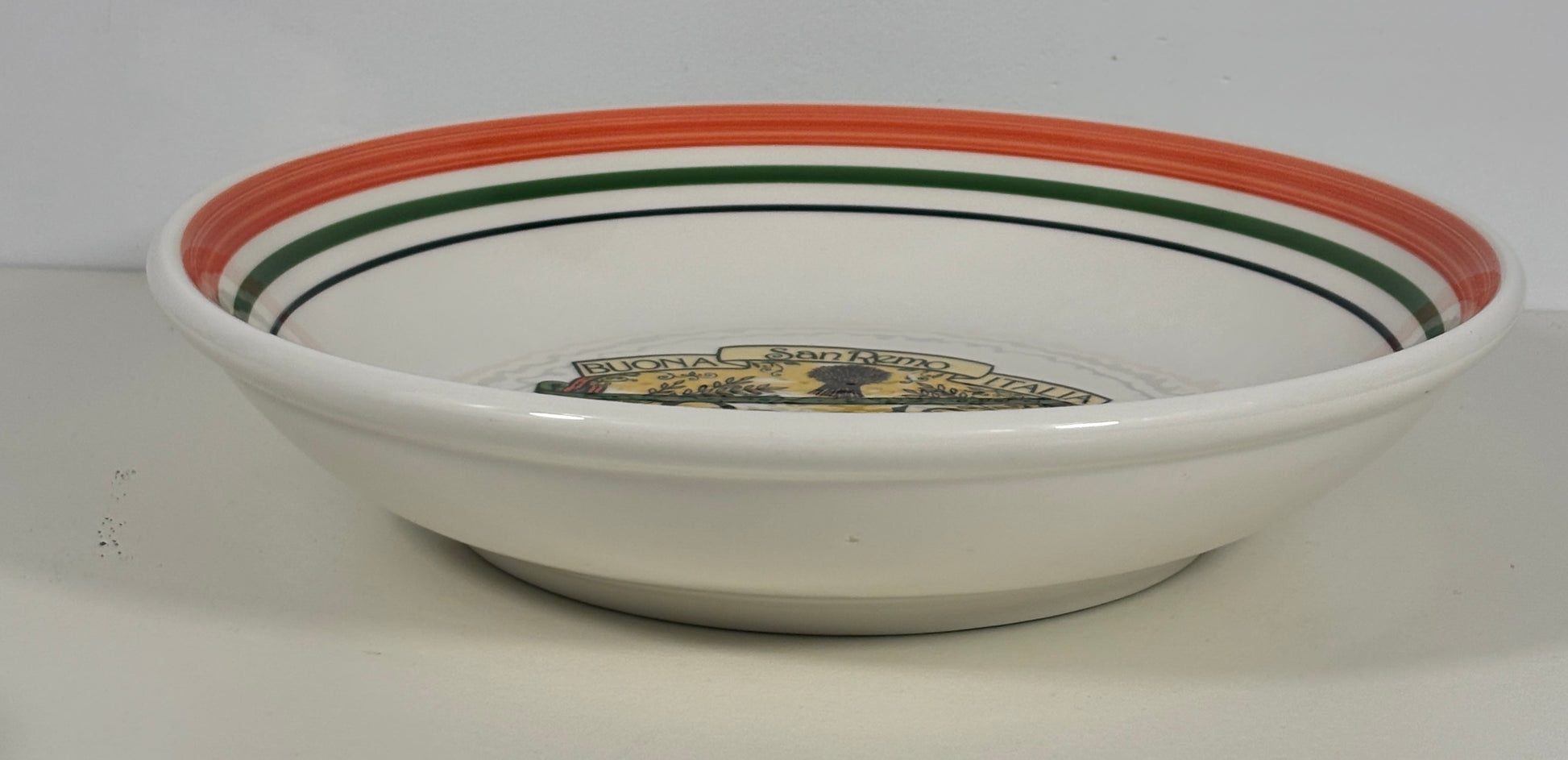 A white ceramic bowl with a wide rim decorated with concentric stripes in orange, green, and black, featuring a small gold and black emblem printed inside the bottom.