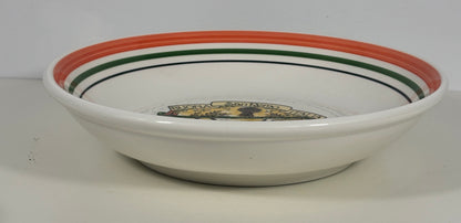 A white ceramic bowl with a wide rim decorated with concentric stripes in orange, green, and black, featuring a small gold and black emblem printed inside the bottom.