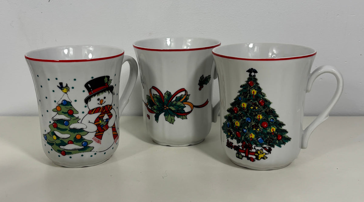 Three white ceramic Christmas mugs with red rims, featuring holiday illustrations including a snowman, a bow, and a decorated Christmas tree.