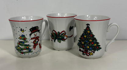 Three white ceramic Christmas mugs with red rims, featuring holiday illustrations including a snowman, a bow, and a decorated Christmas tree.