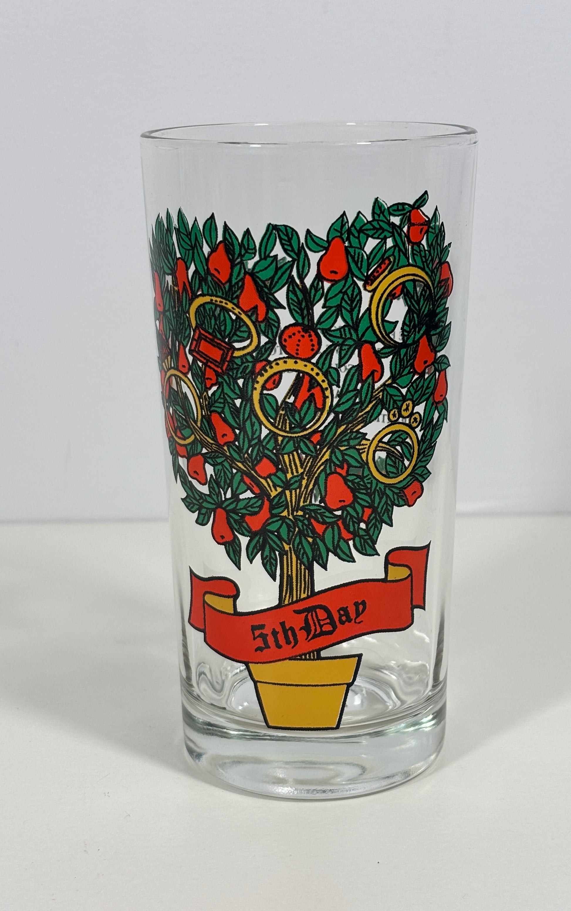 A clear glass tumbler featuring a vintage-style illustration of a tree with red apples, green leaves, gold rings, and a red banner reading '5th May'.