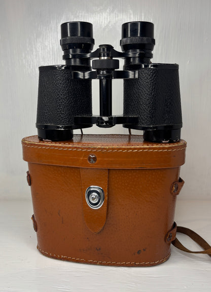 Vintage binoculars in a brown leather case on a light wooden surface.