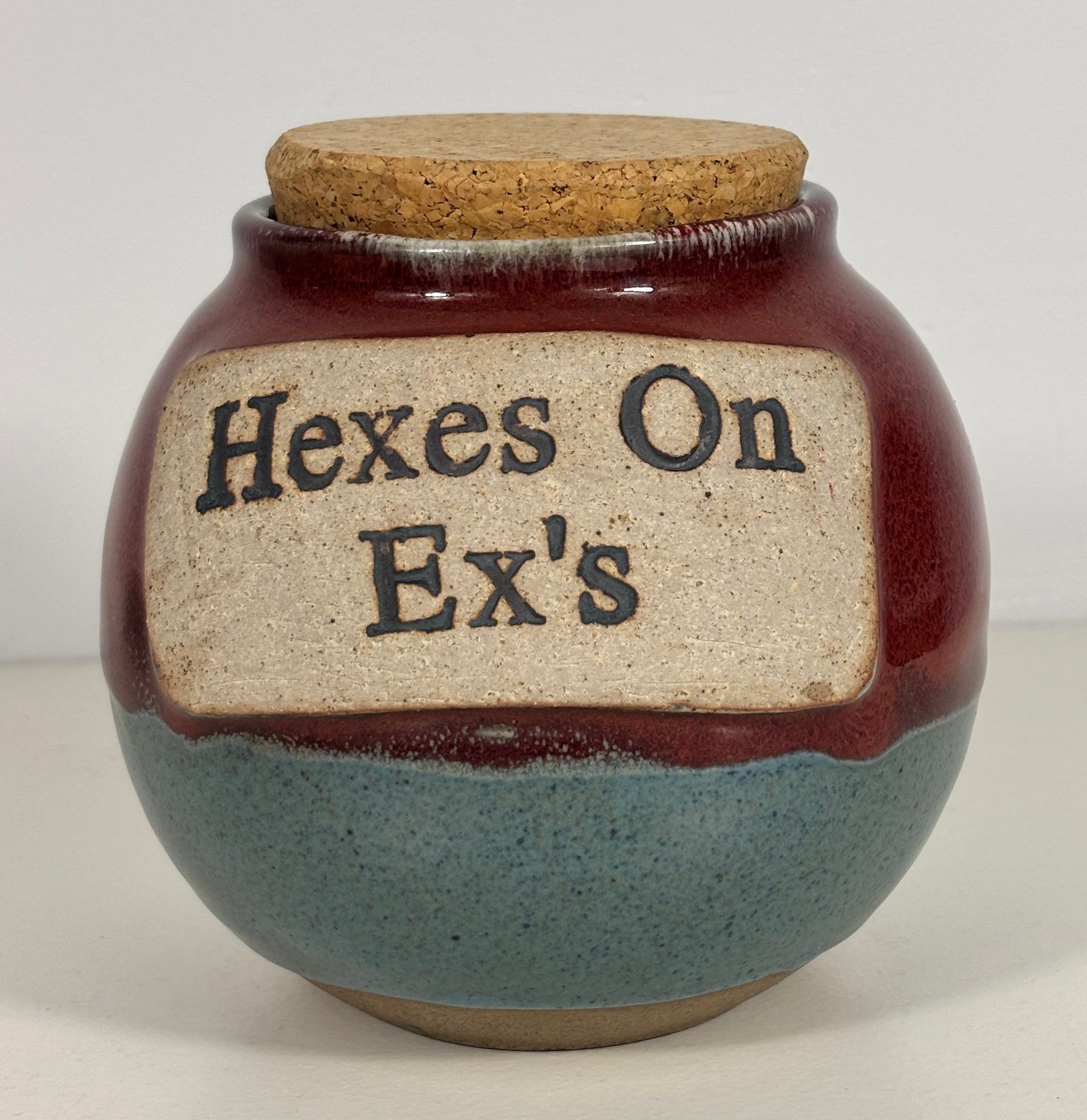 A handmade ceramic jar with a cork lid, featuring a dark reddish-brown glaze on the upper half, a speckled slate-blue glaze on the lower half, and a beige label with the text 'Hexes On Ex's' in black.