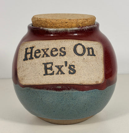A handmade ceramic jar with a cork lid, featuring a dark reddish-brown glaze on the upper half, a speckled slate-blue glaze on the lower half, and a beige label with the text 'Hexes On Ex's' in black.