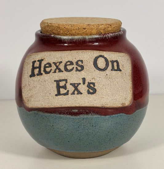 A handmade ceramic jar with a cork lid, featuring a dark reddish-brown glaze on the upper half, a speckled slate-blue glaze on the lower half, and a beige label with the text 'Hexes On Ex's' in black.