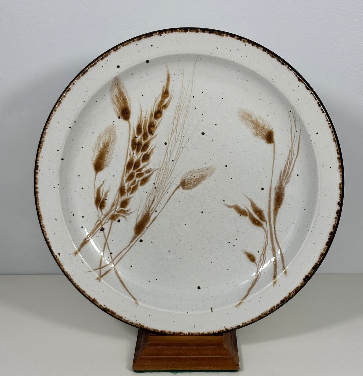 A round ceramic plate with a speckled off-white glaze, decorated with brown painted wheat stalks and displayed on a small wooden stand against a plain light gray background.