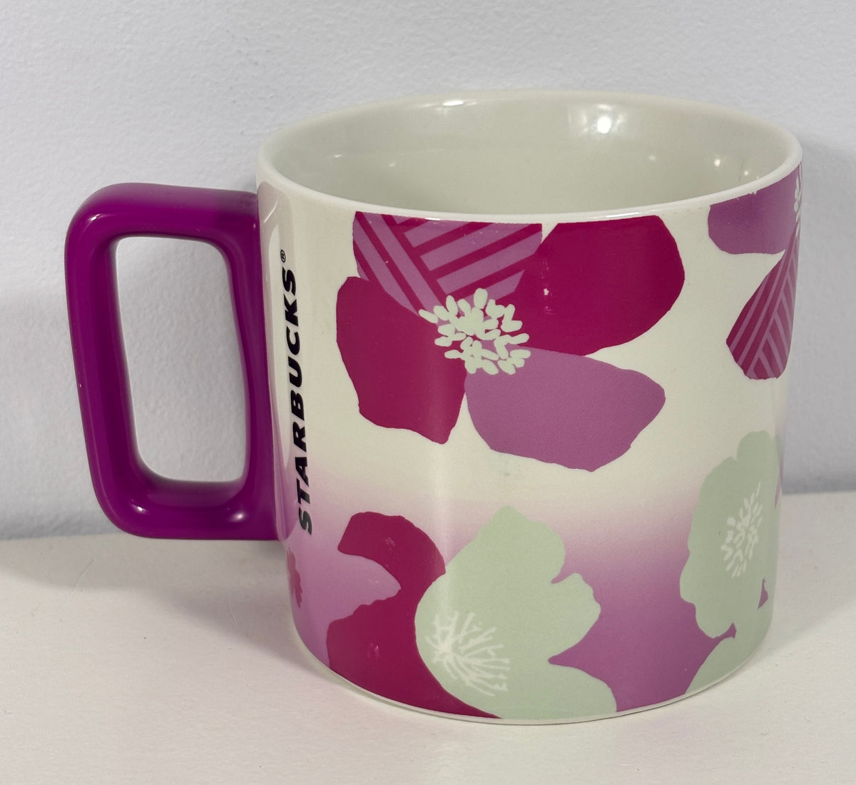 A Starbucks ceramic mug with a purple handle, featuring a floral pattern in shades of magenta, purple, and light green on a white background.