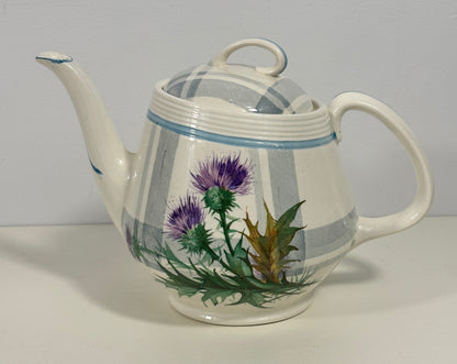 A vintage ceramic teapot with a white body, gray and blue plaid pattern, and a hand-painted purple thistle and green leaves design on the front, set against a plain light gray background.