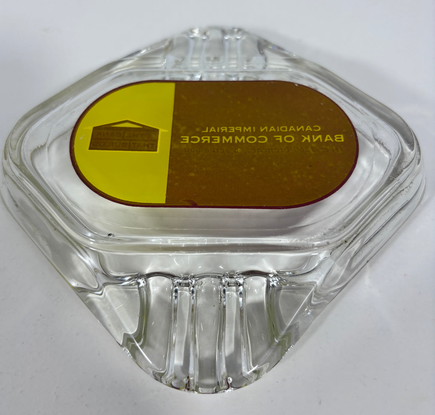 A clear glass ashtray with a hexagonal shape and a circular gold and brown label on top that reads 'BANK OF COMMERCE CANADIAN IMPERIAL'.