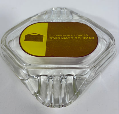 A clear glass ashtray with a hexagonal shape and a circular gold and brown label on top that reads 'BANK OF COMMERCE CANADIAN IMPERIAL'.