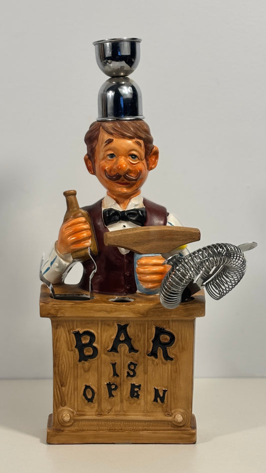 A novelty bar-themed light holder shaped like a bartender figurine standing behind a wooden counter with the words 'BAR IS OPEN' on the front. The figurine holds a bottle and a metal tin, with a small metallic cup balanced on its head.