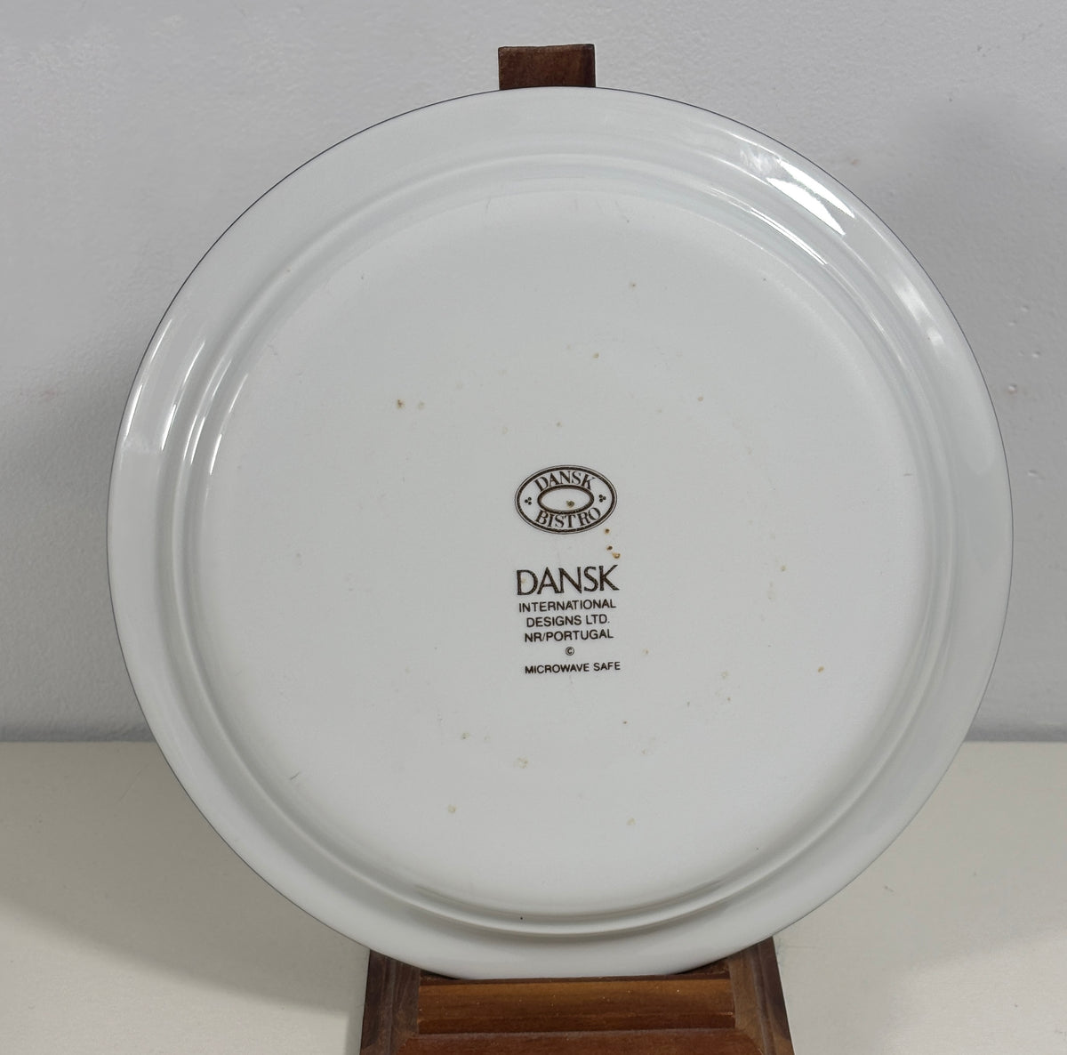 A white ceramic plate with a black printed Dansk International logo in the center, displayed on a wooden stand against a plain light gray background.