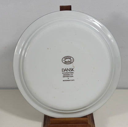 A white ceramic plate with a black printed Dansk International logo in the center, displayed on a wooden stand against a plain light gray background.