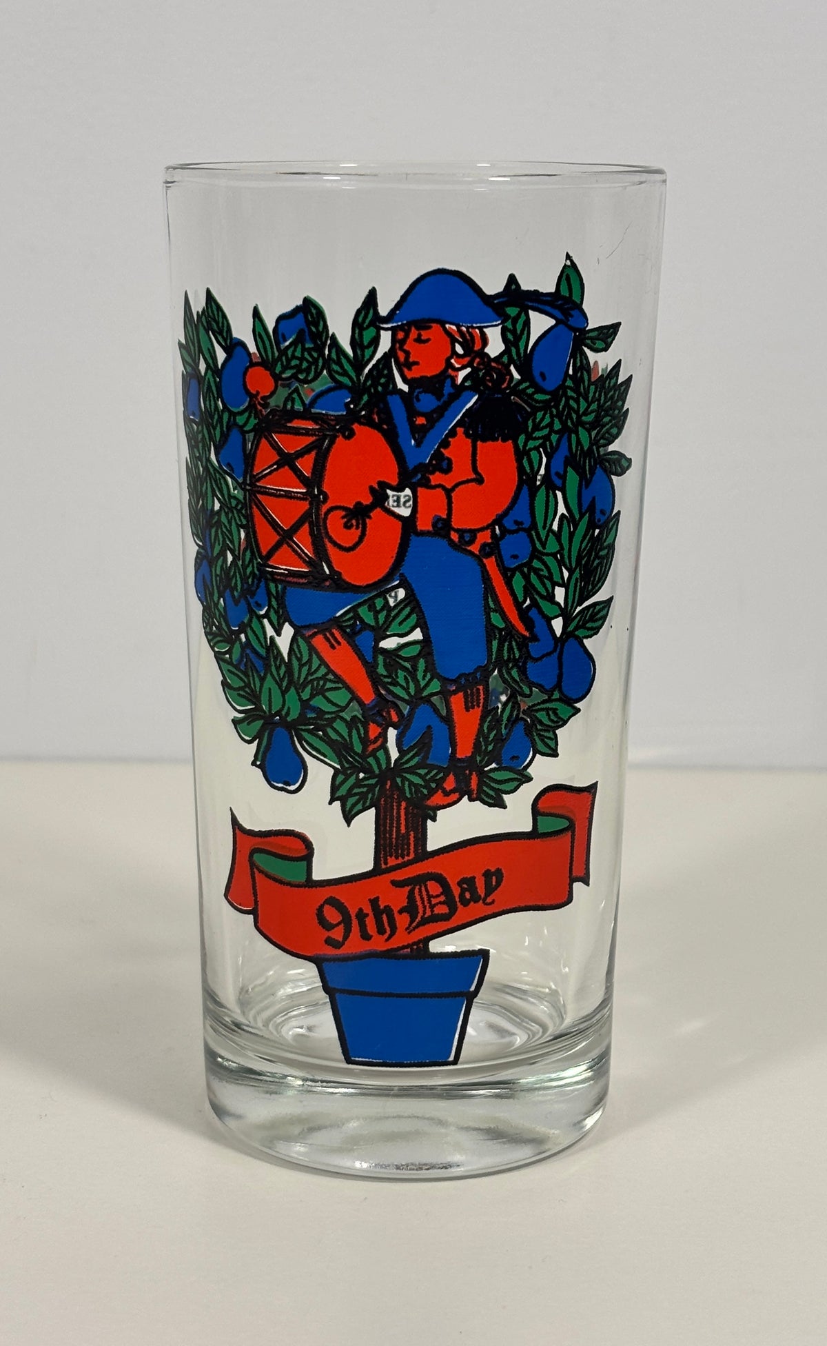 A clear glass tumbler featuring a colorful printed graphic of a soldier playing a drum surrounded by blue berries, with a red banner reading '9th Bar' at the bottom, set against a plain light background.