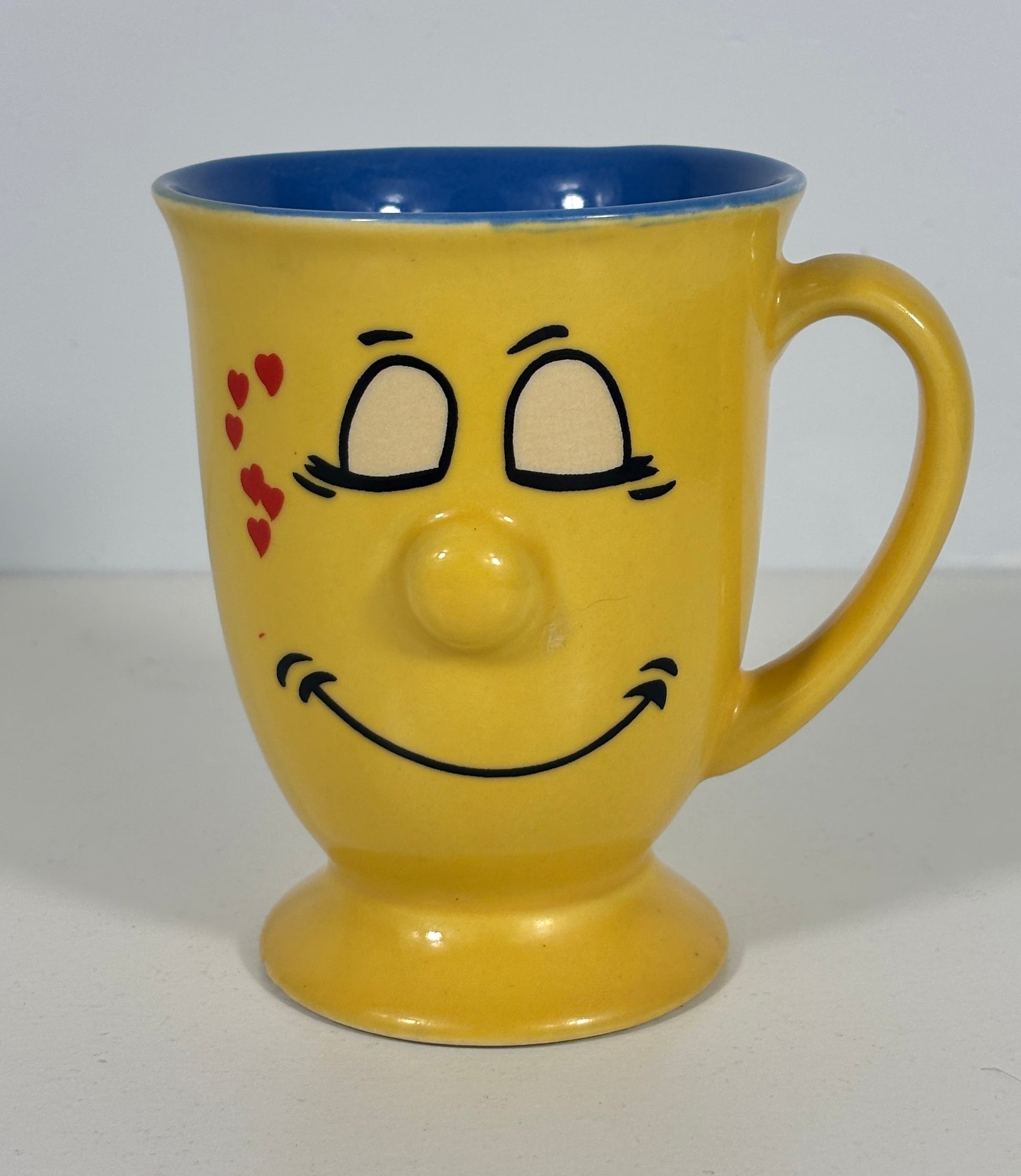 A yellow ceramic mug with a blue interior, featuring a hand-painted smiling face with red hearts on the cheek.