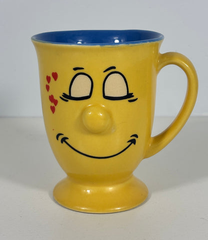 A yellow ceramic mug with a blue interior, featuring a hand-painted smiling face with red hearts on the cheek.