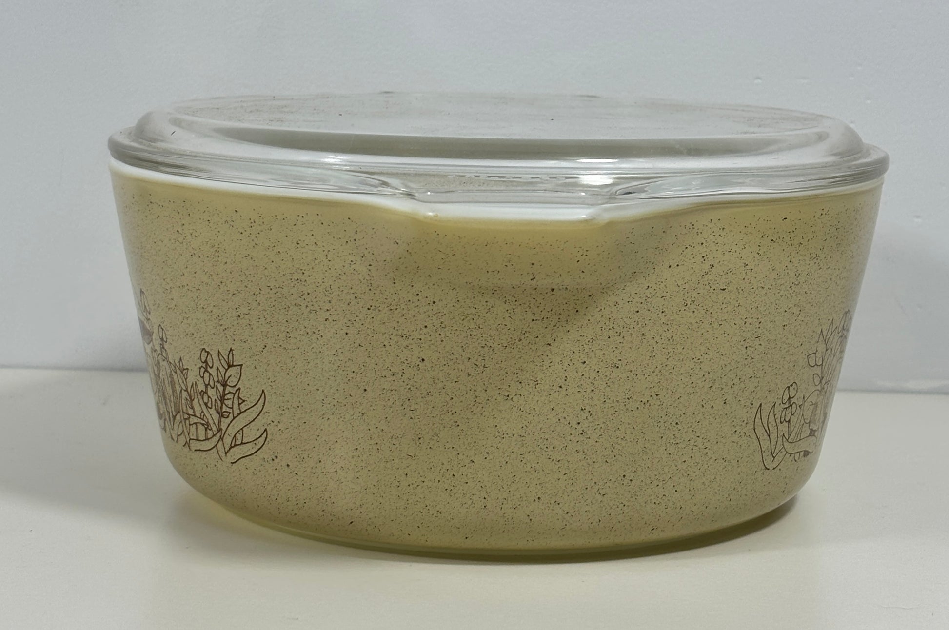 A vintage Pyrex glass casserole dish with a clear glass lid, featuring a speckled beige body with etched floral designs on the sides.