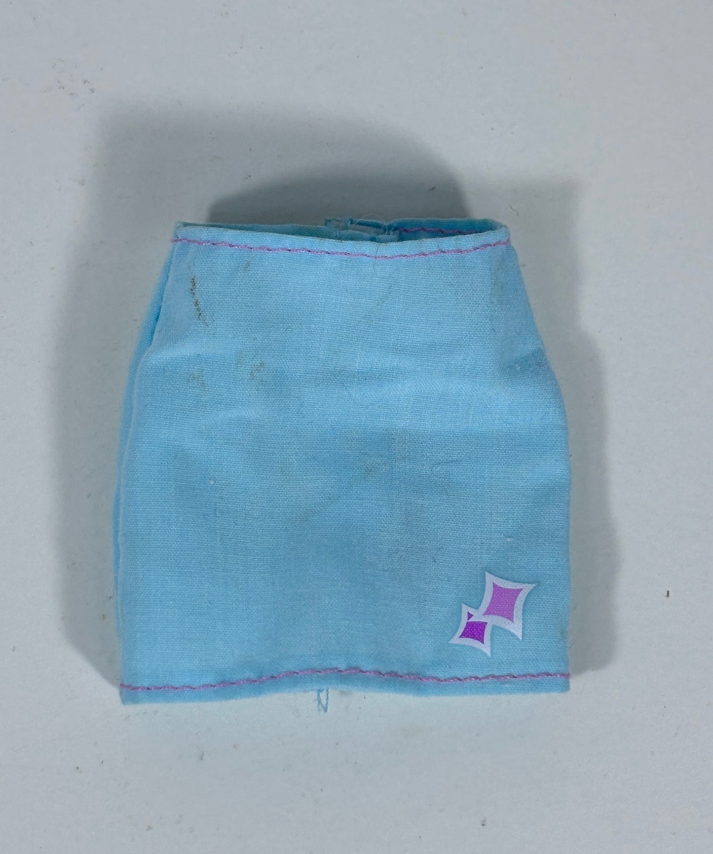 A light blue fabric skirt or shorts with pink stitching and a small purple and white starburst logo on the lower right, photographed against a plain light gray background.