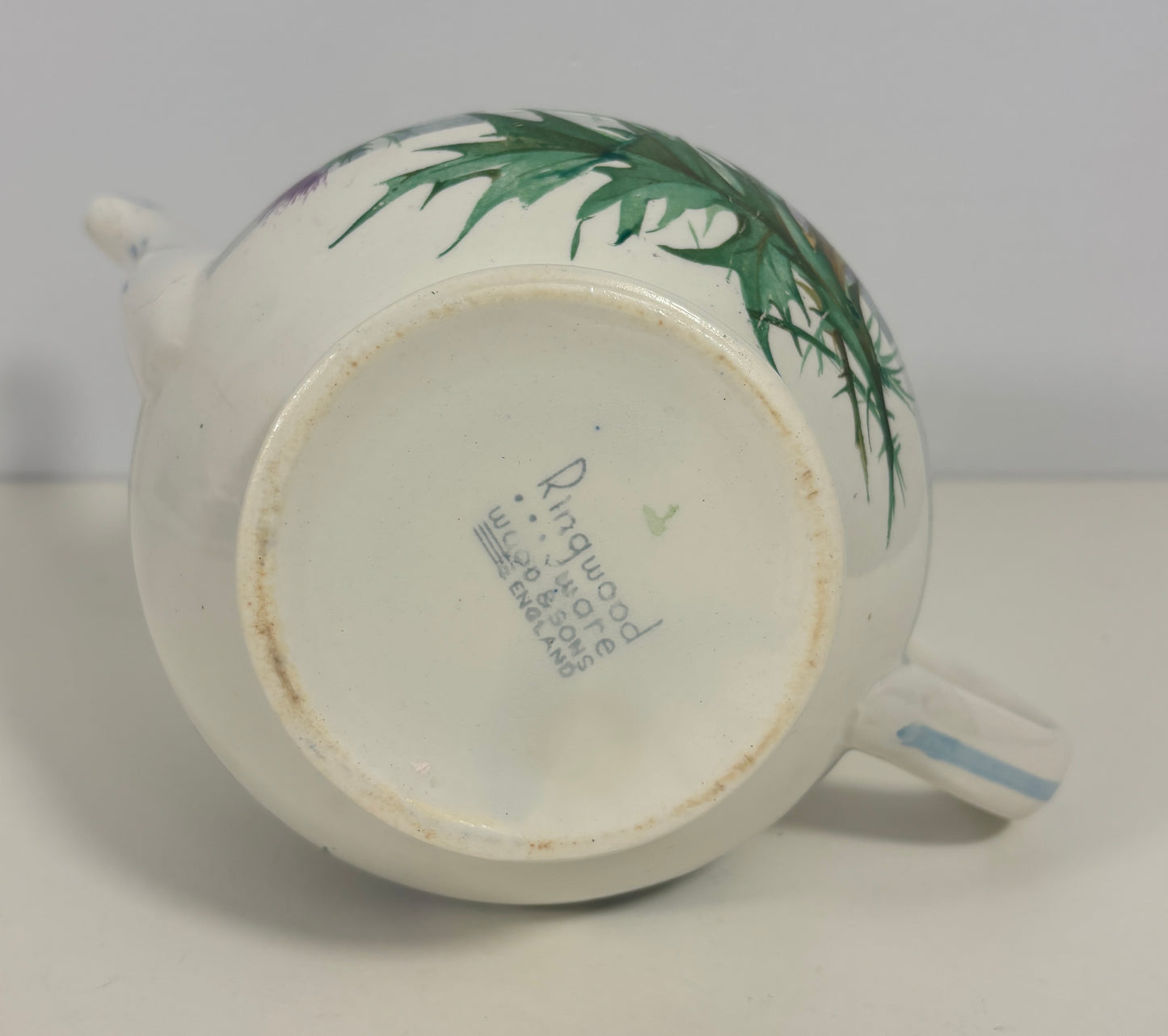 A vintage white porcelain teapot with hand-painted green leaves on the side, shown from a bottom-up angle to display the maker's mark 'Rugwood' and 'No. 7' on the base.