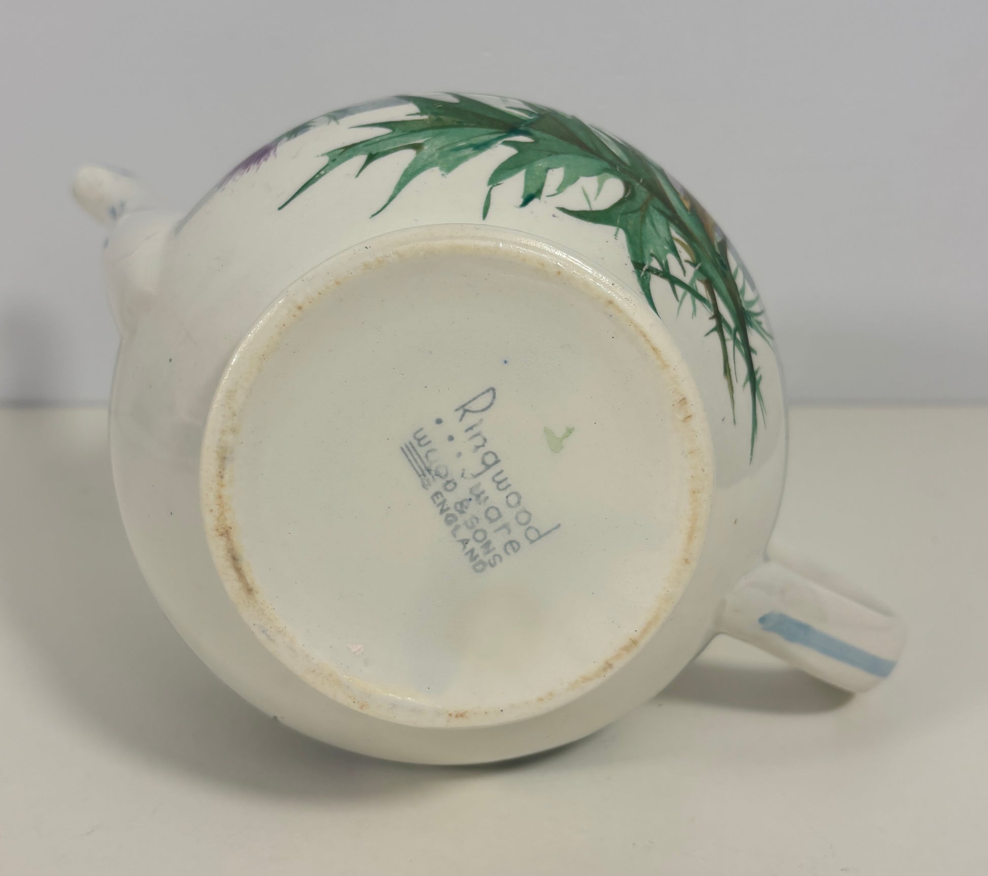 A vintage white porcelain teapot with hand-painted green leaves on the side, shown from a bottom-up angle to display the maker's mark 'Rugwood' and 'No. 7' on the base.
