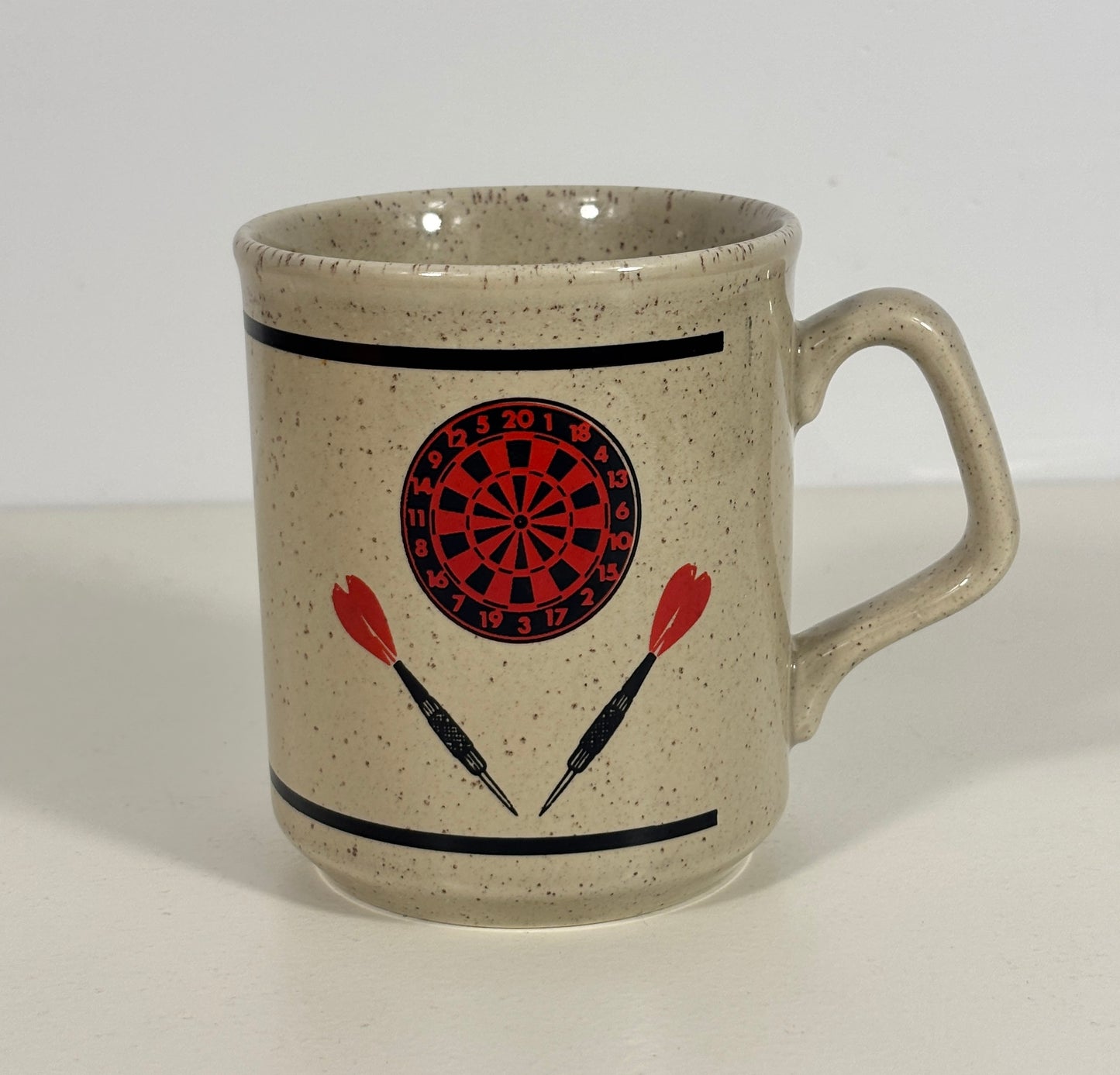 A speckled beige ceramic mug with a darts theme, featuring a red and black dartboard graphic and two red-and-black darts printed on the front, set against a plain white background.