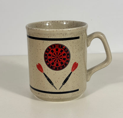 A speckled beige ceramic mug with a darts theme, featuring a red and black dartboard graphic and two red-and-black darts printed on the front, set against a plain white background.