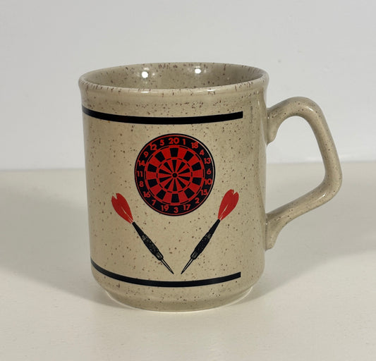 A speckled beige ceramic mug with a darts theme, featuring a red and black dartboard graphic and two red-and-black darts printed on the front, set against a plain white background.