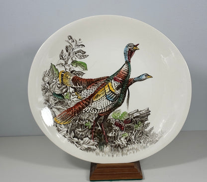 A round white ceramic plate featuring a detailed illustration of two wild turkeys in natural foliage, displayed on a small wooden stand against a neutral gray background.