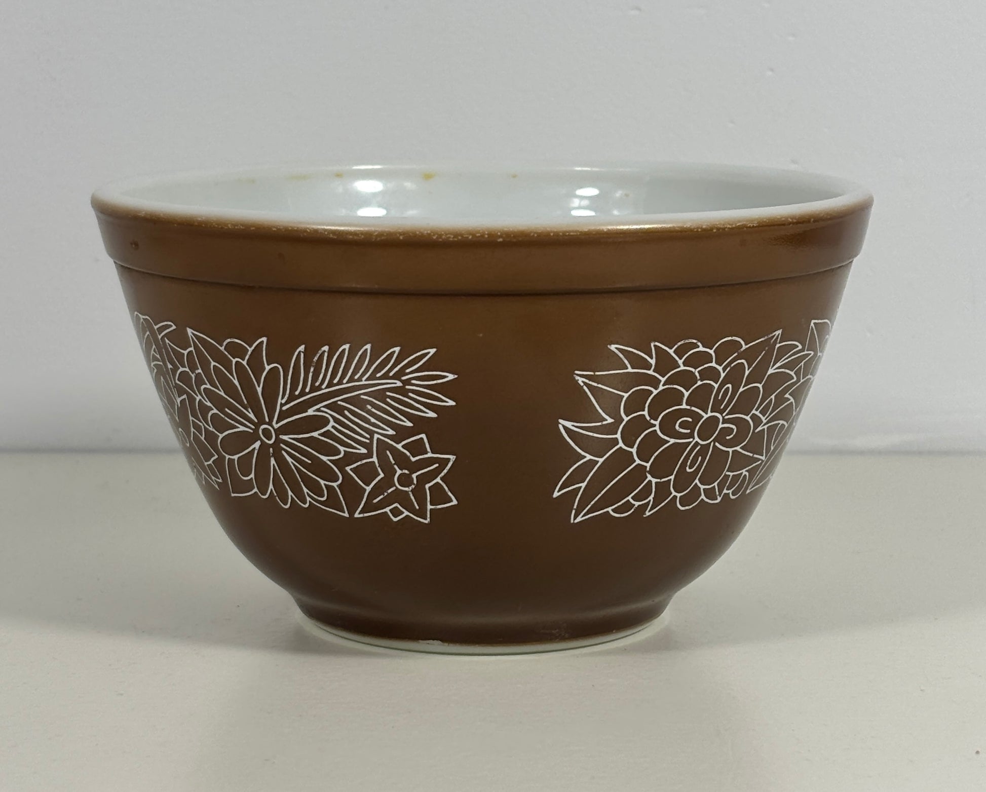 A brown mixing bowl with a white interior and white line-art floral pattern on the exterior, set against a plain light gray background.