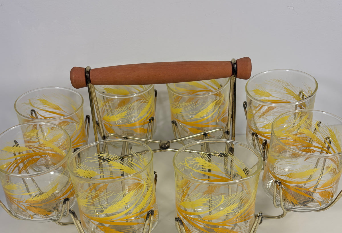 A set of 8 clear glass tumblers with yellow feather designs, arranged around a metal and wood carrier with a brown wooden handle, on a plain white background.