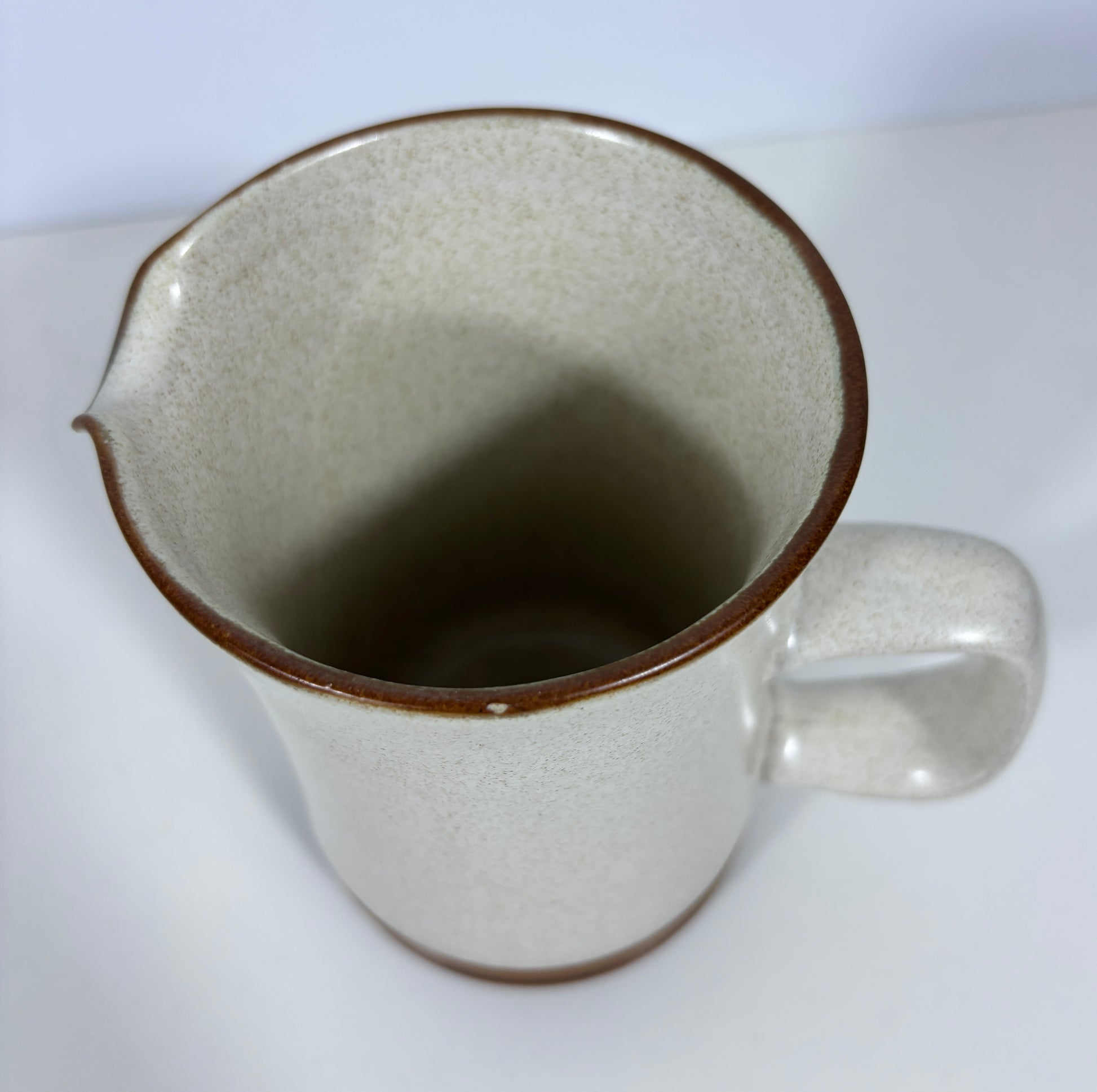 A speckled ceramic pitcher with a warm off-white glaze and a contrasting dark brown rim and base, shown on a plain white surface.