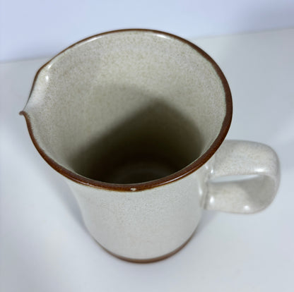 A speckled ceramic pitcher with a warm off-white glaze and a contrasting dark brown rim and base, shown on a plain white surface.